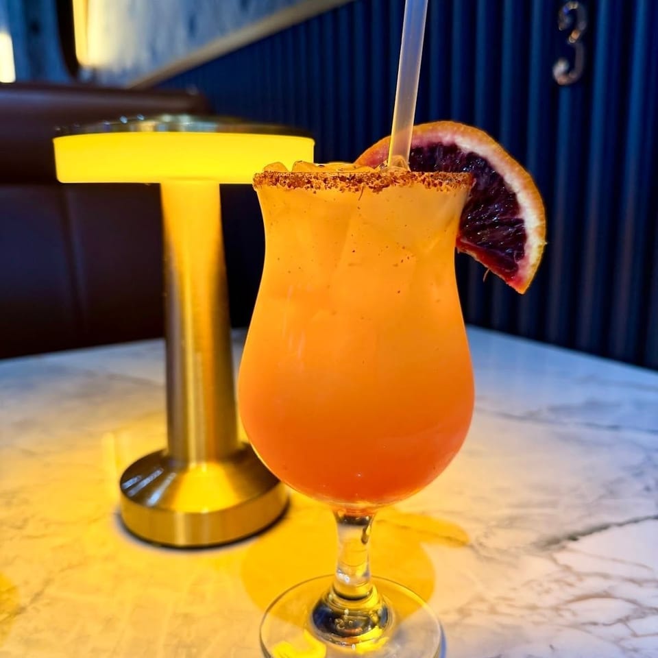 Orange cocktail with a chili-salt rim, straw, and blood orange garnish on a marble table beside a glowing lamp.