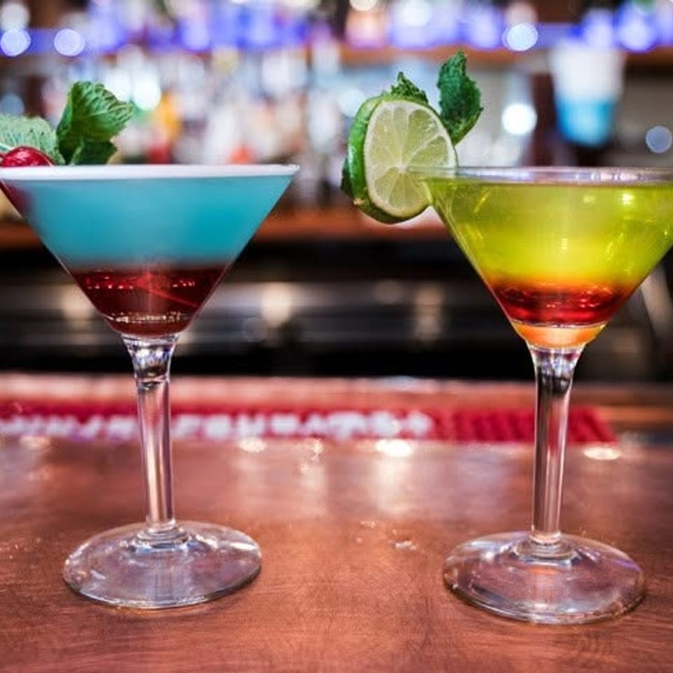 Two colorful layered cocktails in martini glasses, garnished with fresh mint, berries, and lime, sit on a bar counter.