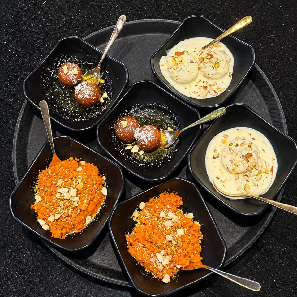 Assorted Indian desserts in black bowls, including gulab jamun, creamy rasmalai, and orange carrot halwa topped with nuts.