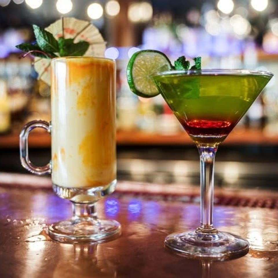Two colorful cocktails sit on a polished bar, including a creamy layered drink and a lime-garnished green martini.