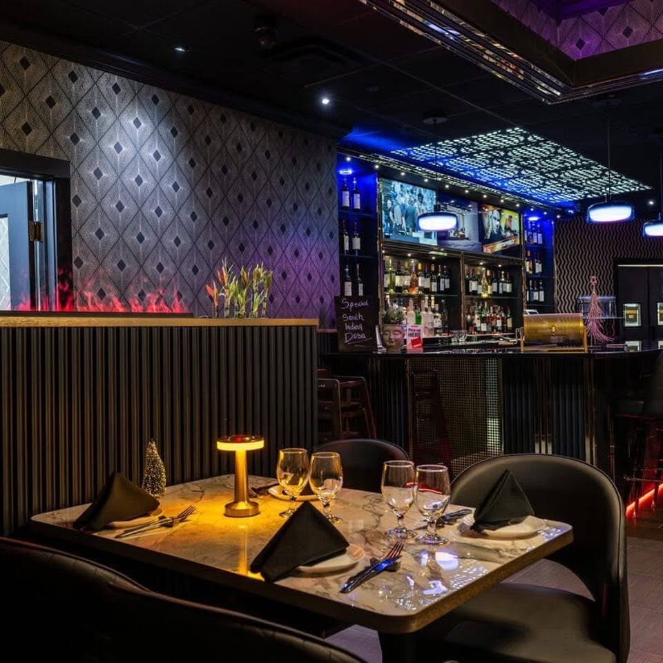 Upscale restaurant interior with a set dining table, marble bar, patterned walls, colorful lighting, and rows of bar stools.