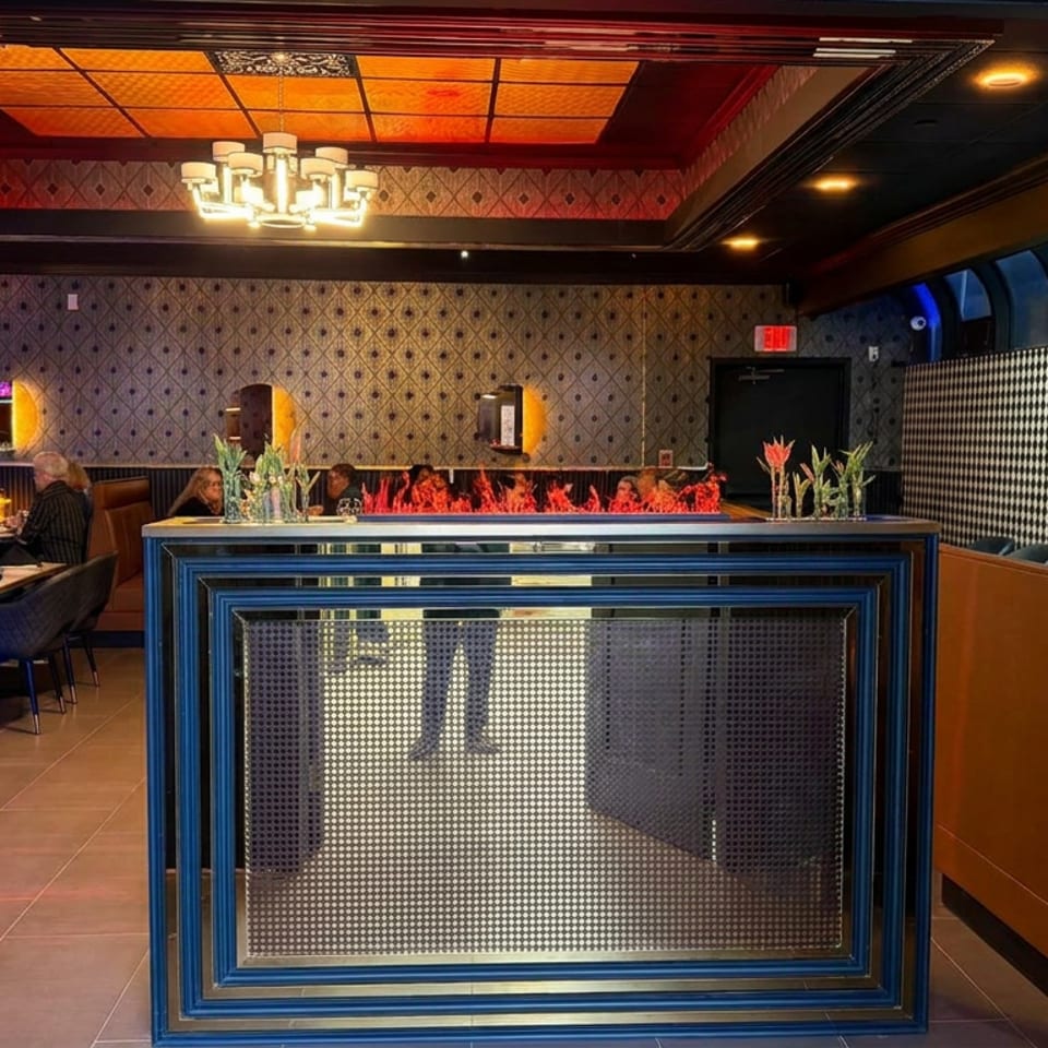 Elegant restaurant interior viewed through blue doors, featuring a tiled bar, glowing fireplace, patterned walls, booths, and chandelier lighting.